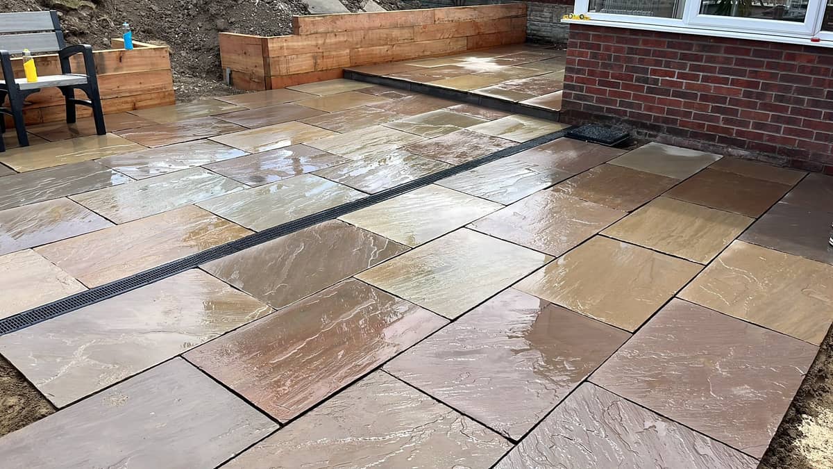 A wide sandstone patio across the back garden, raised timber beds at the far end